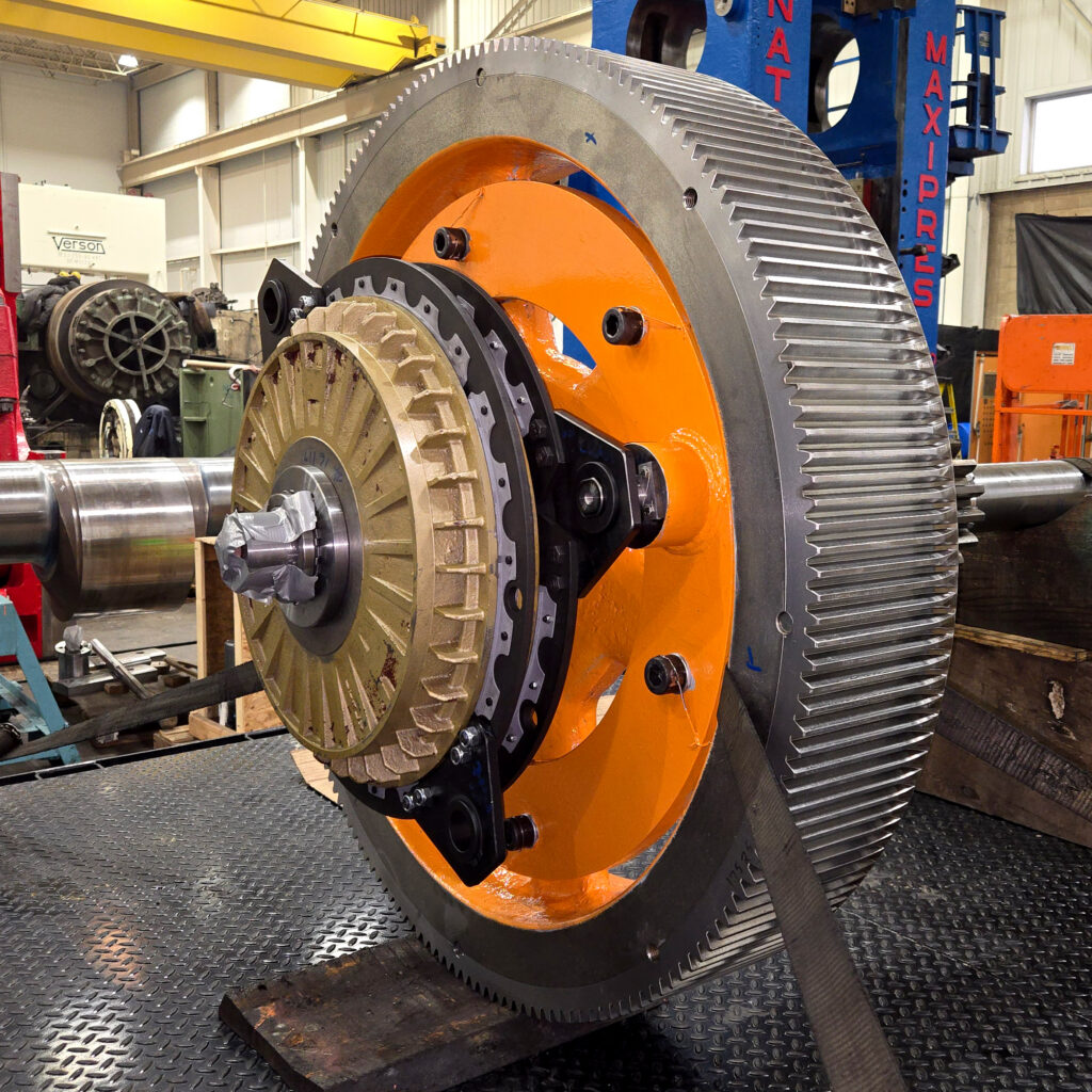 Completed industrial clutch conversion featuring a large machined flywheel, bronze clutch assembly, and orange mounting plate installed on a heavy-duty shaft in a machine shop.
