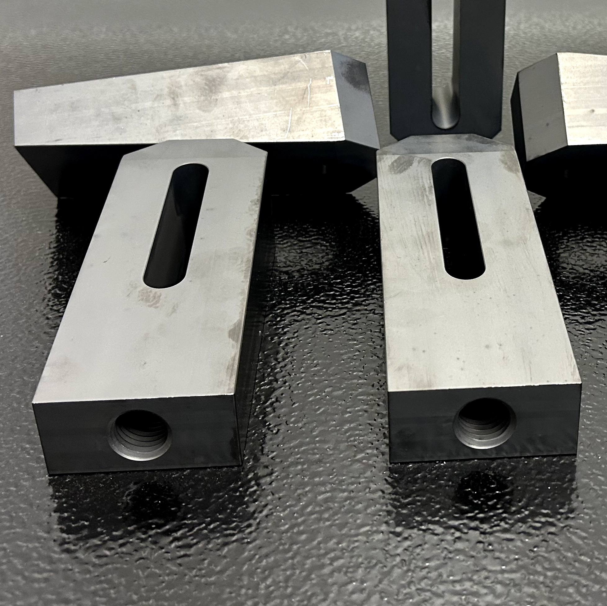 Close-up of two precision-machined wedge blocks with elongated mounting slots and threaded end holes, displayed on a black textured surface.