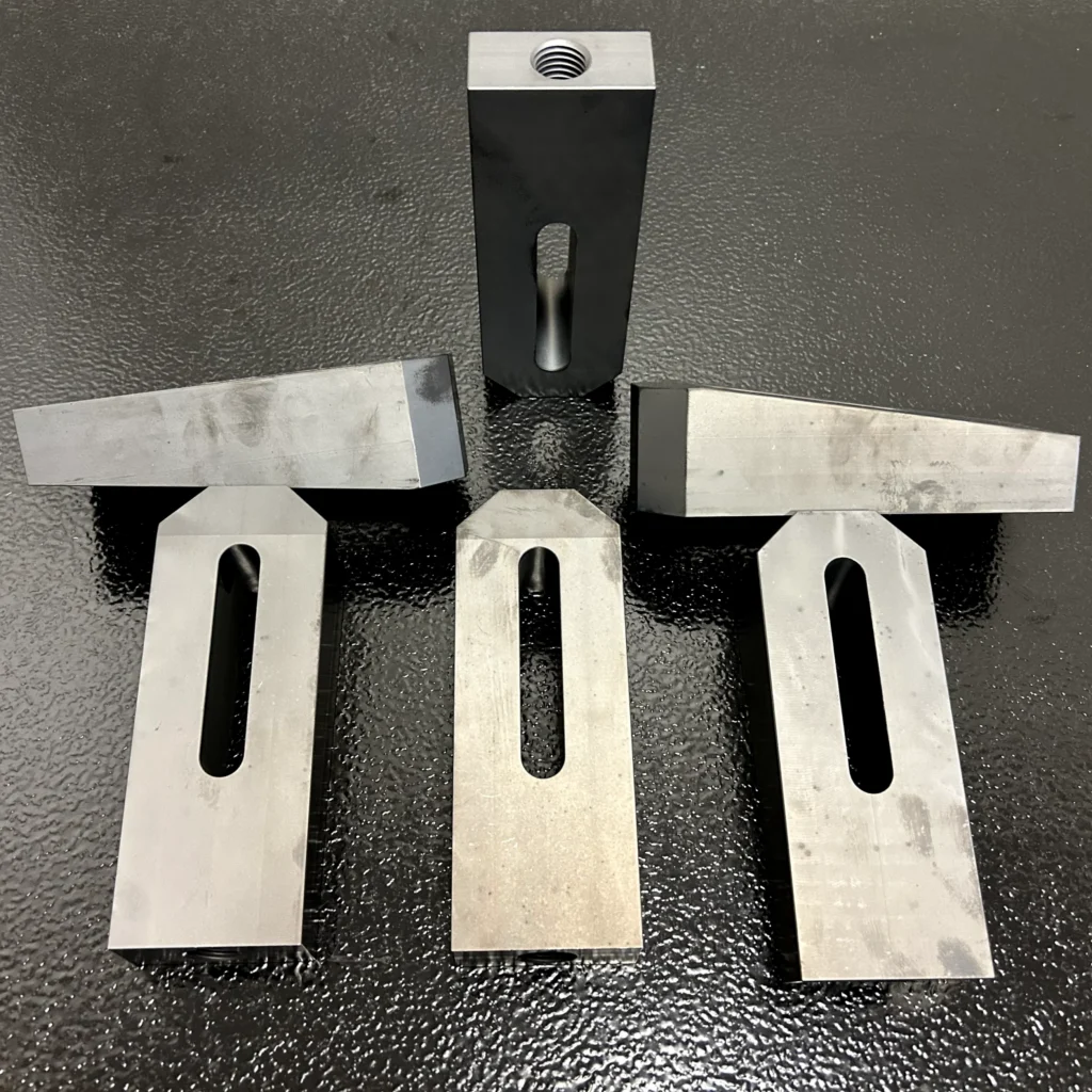 Six precision-machined wedge blocks shown from multiple angles, featuring tapered wedge tops, elongated slots, and threaded mounting holes on a black textured surface.