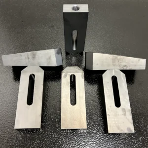 Six precision-machined wedge blocks shown from multiple angles, featuring tapered wedge tops, elongated slots, and threaded mounting holes on a black textured surface.