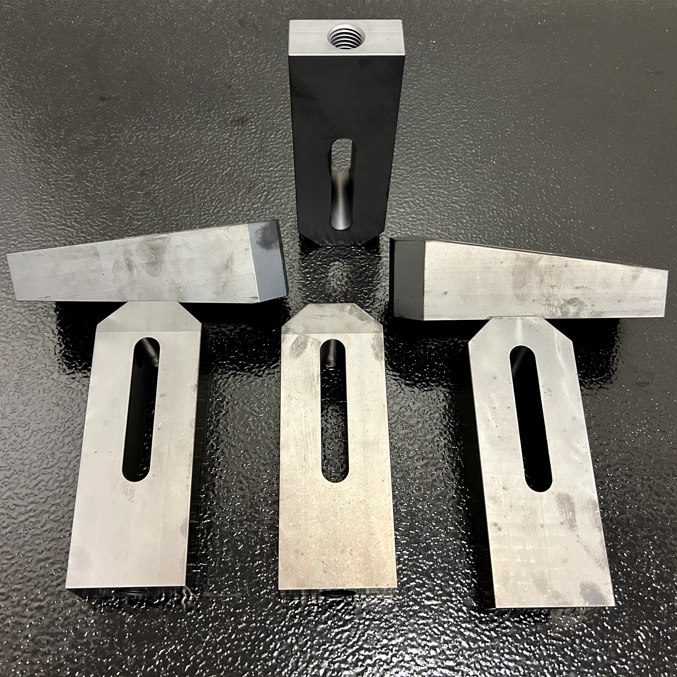 Six precision-machined wedge blocks shown from multiple angles, featuring tapered wedge tops, elongated slots, and threaded mounting holes on a black textured surface.
